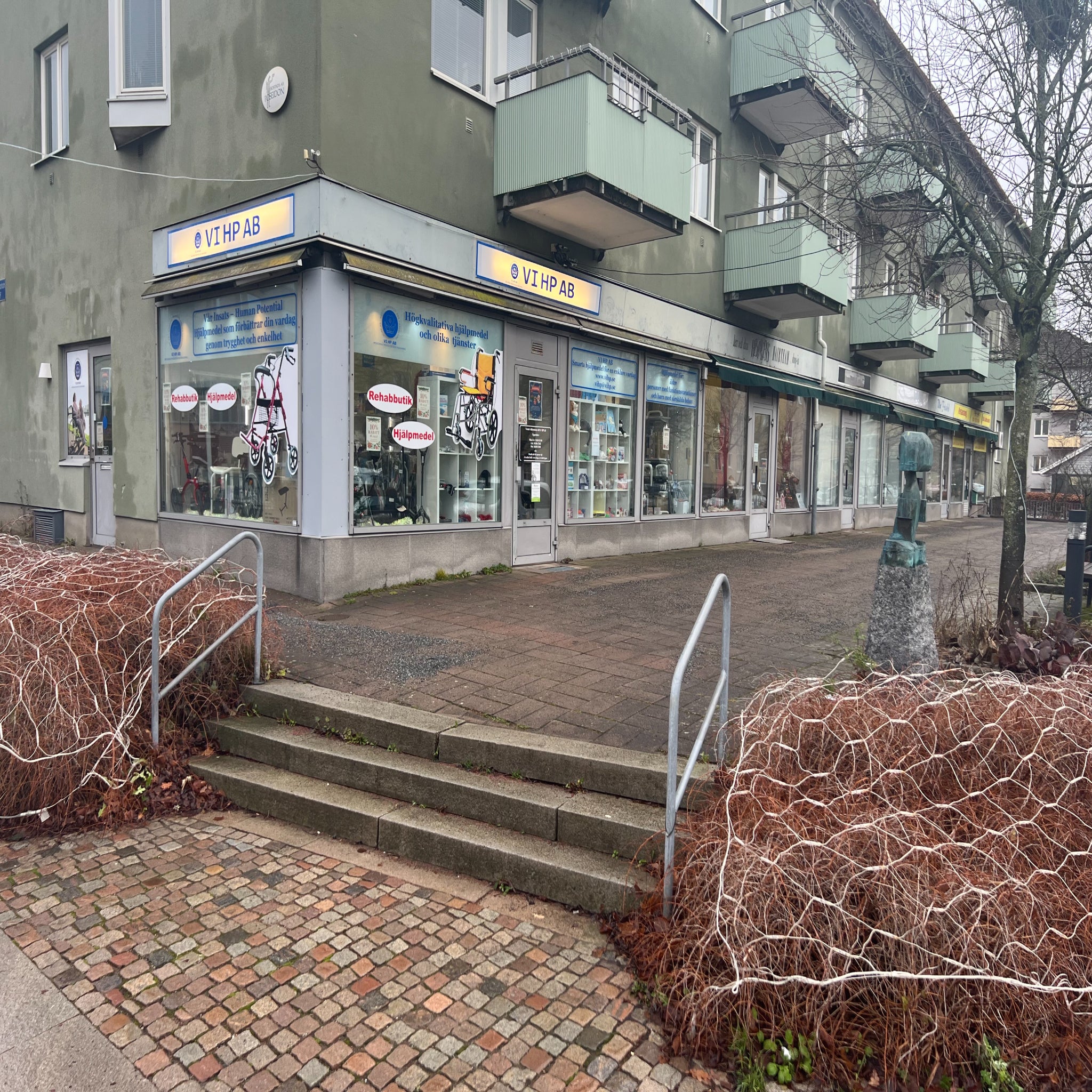 Hjälpmedel och Rehabbutik VI HP fasad med skyltfönster och entré vid gångväg och trappsteg utanför butiken på Byalagsgatan.