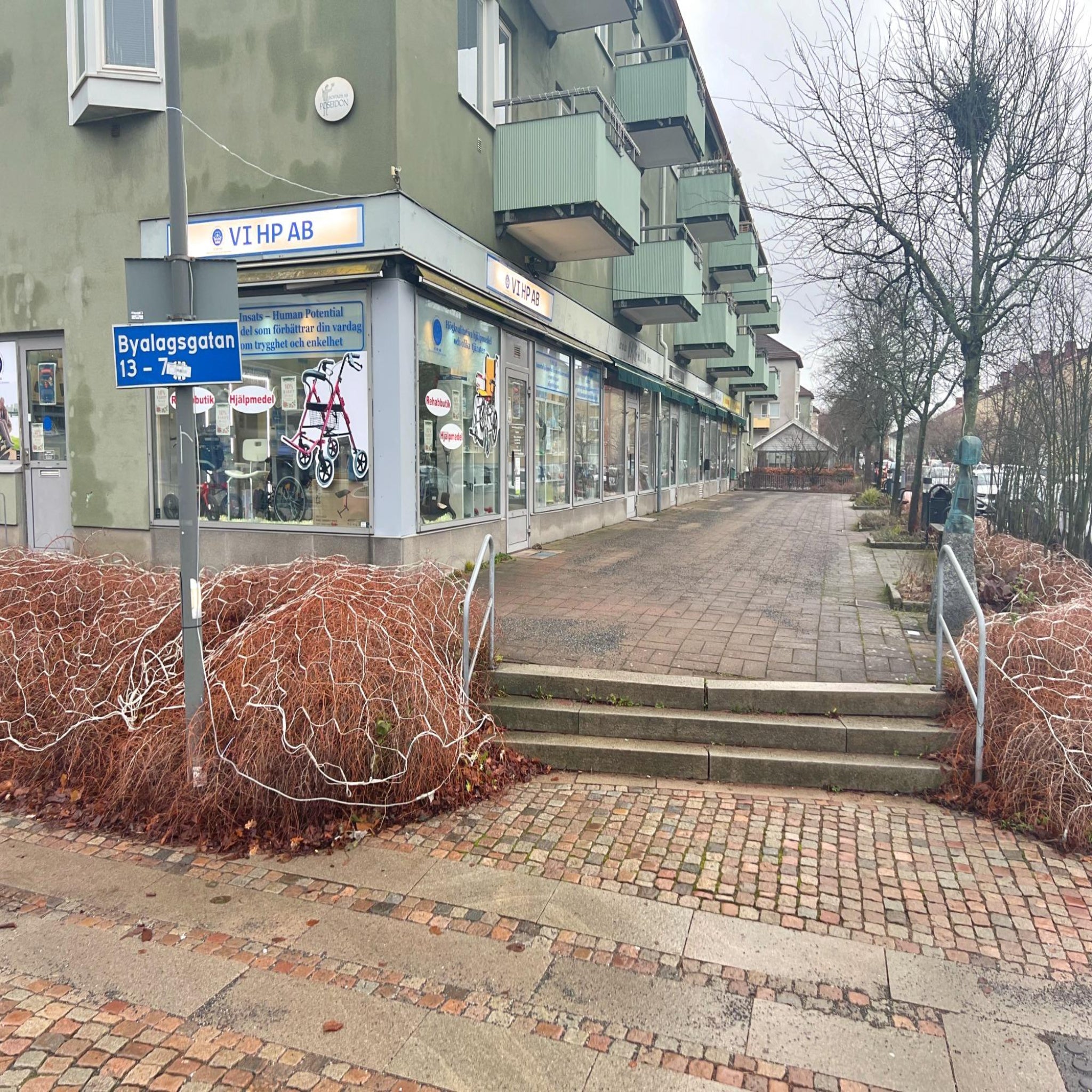 Hjälpmedel och Rehabbutik VI HP fasad vid Byalagsgatan med skyltfönster och entré längs trottoar och gångväg.